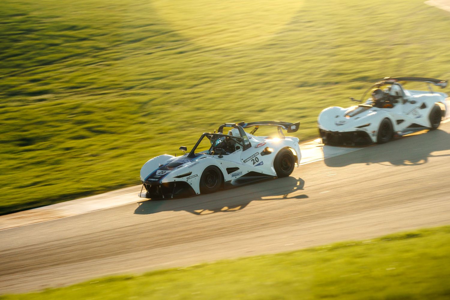 Silver sports cars race around a green racetrack during a competitive motorsport event at sunset.