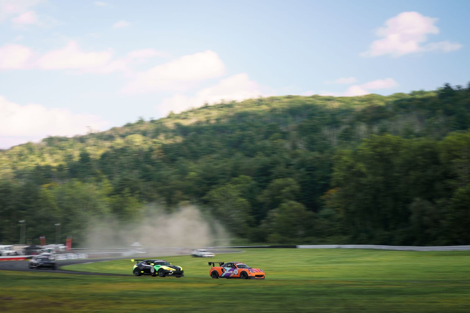 Two race cars speed around a corner on a lush green racetrack surrounded by wooded hills on a sunny day.