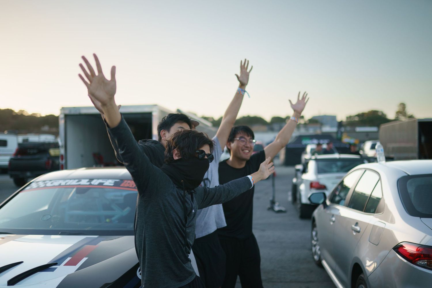A group of people raise their hands in celebration while standing in a parking lot at sunset.