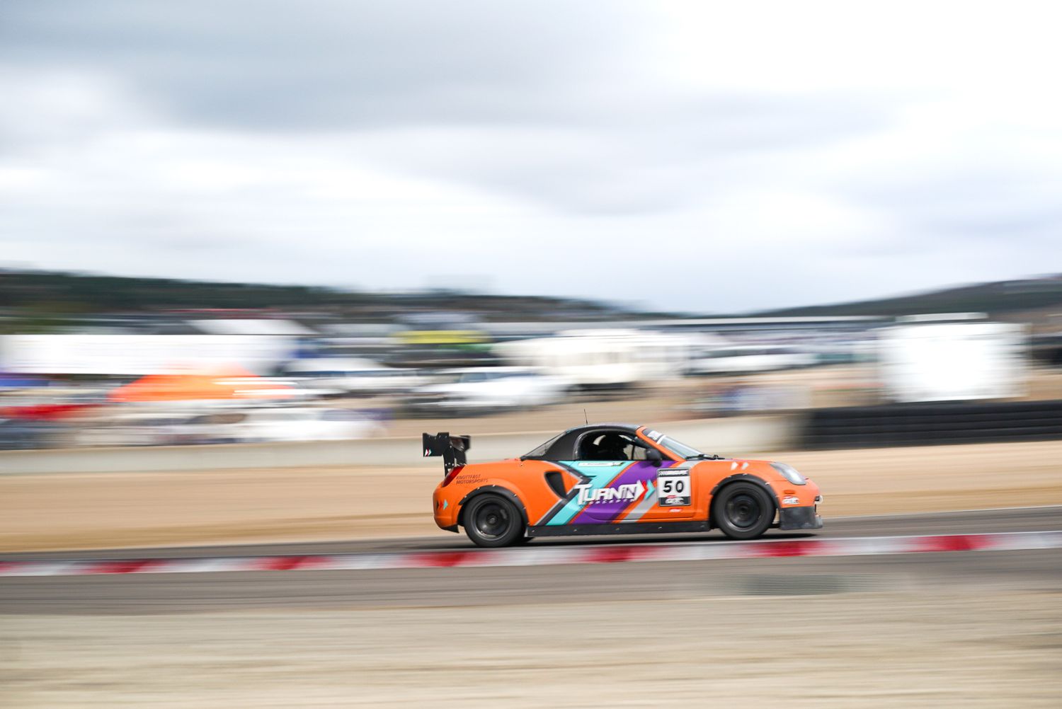 A race car speeds past with motion blur on a track, featuring bright orange and purple livery under cloudy skies.