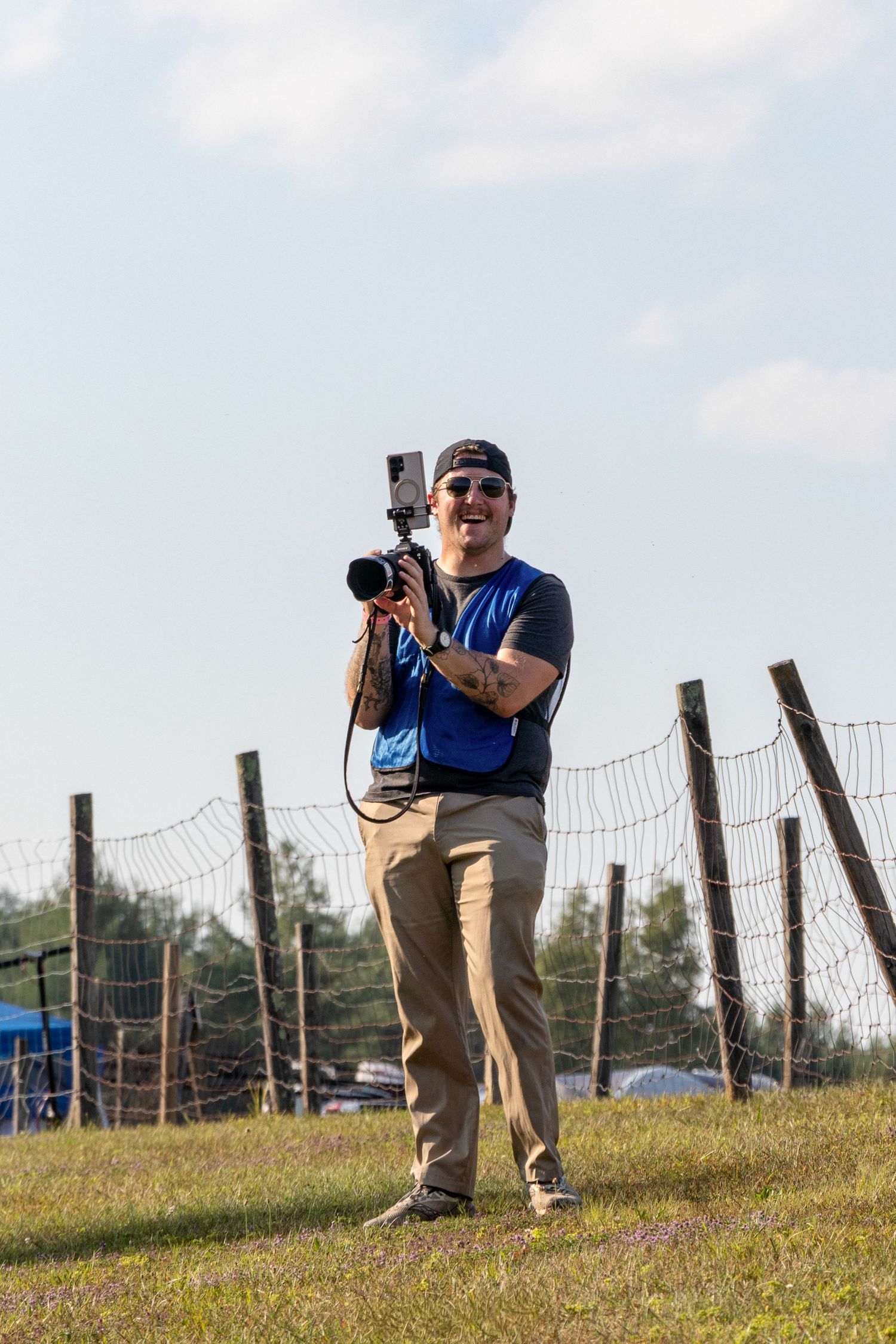 A photographer with a camera takes multiple shots from a low position on a grassy knoll behind fence posts.