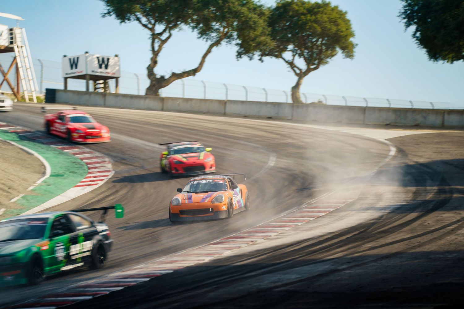 Multiple race cars navigate a challenging turn on a professional racing circuit with trees in the background.