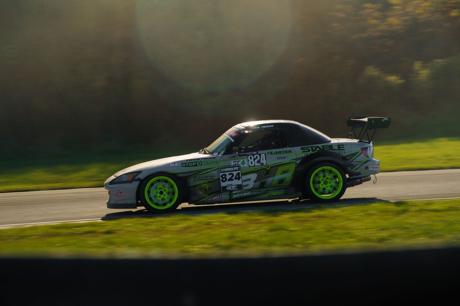 A silver and green racing car speeds along a racetrack during golden hour lighting.