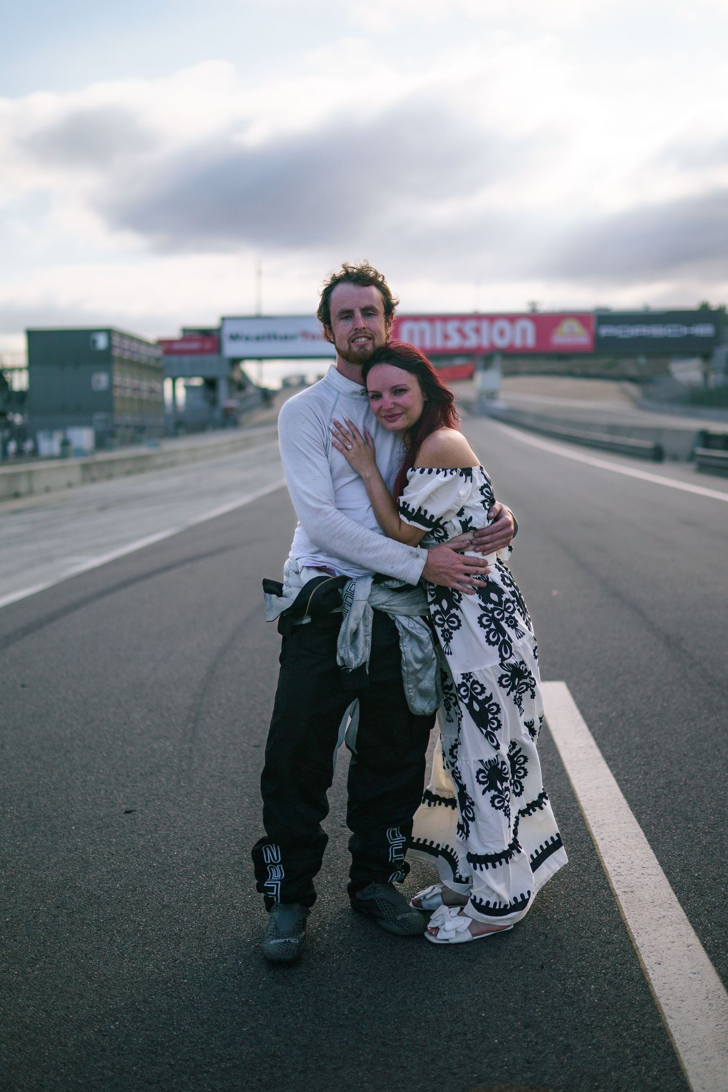 Two people embrace while sitting on an empty racetrack under cloudy skies.
