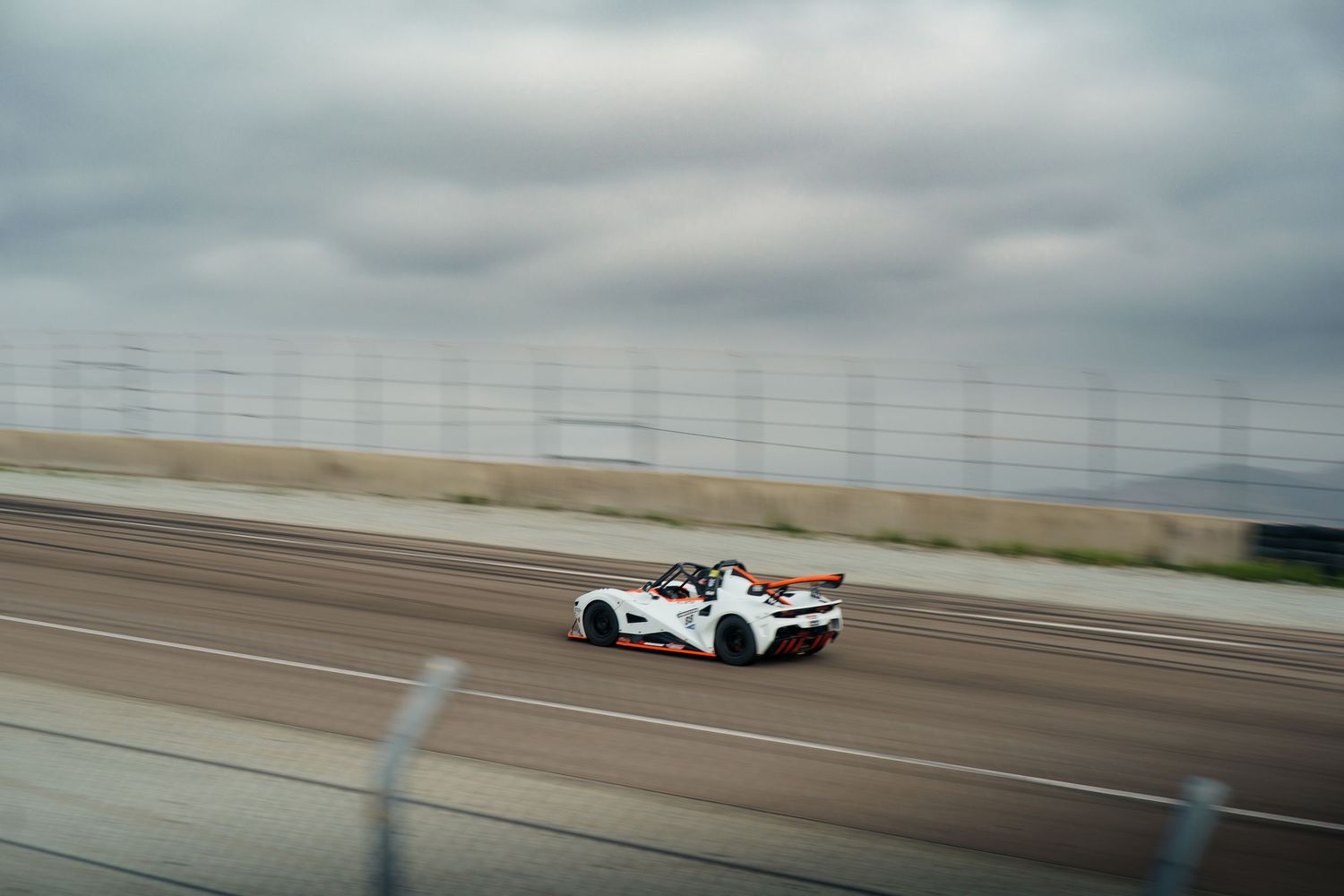White racing car speeds along a racetrack with blurred motion against an overcast sky.