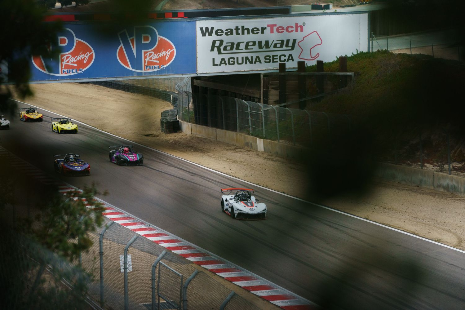 Race cars speed down a track beneath a WeatherTech racing banner during a motorsport competition.