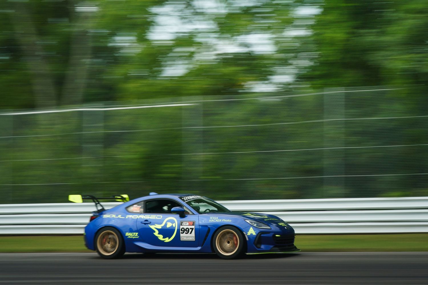 A blue sports car races on a track with green trees in the background during a motorsport event.