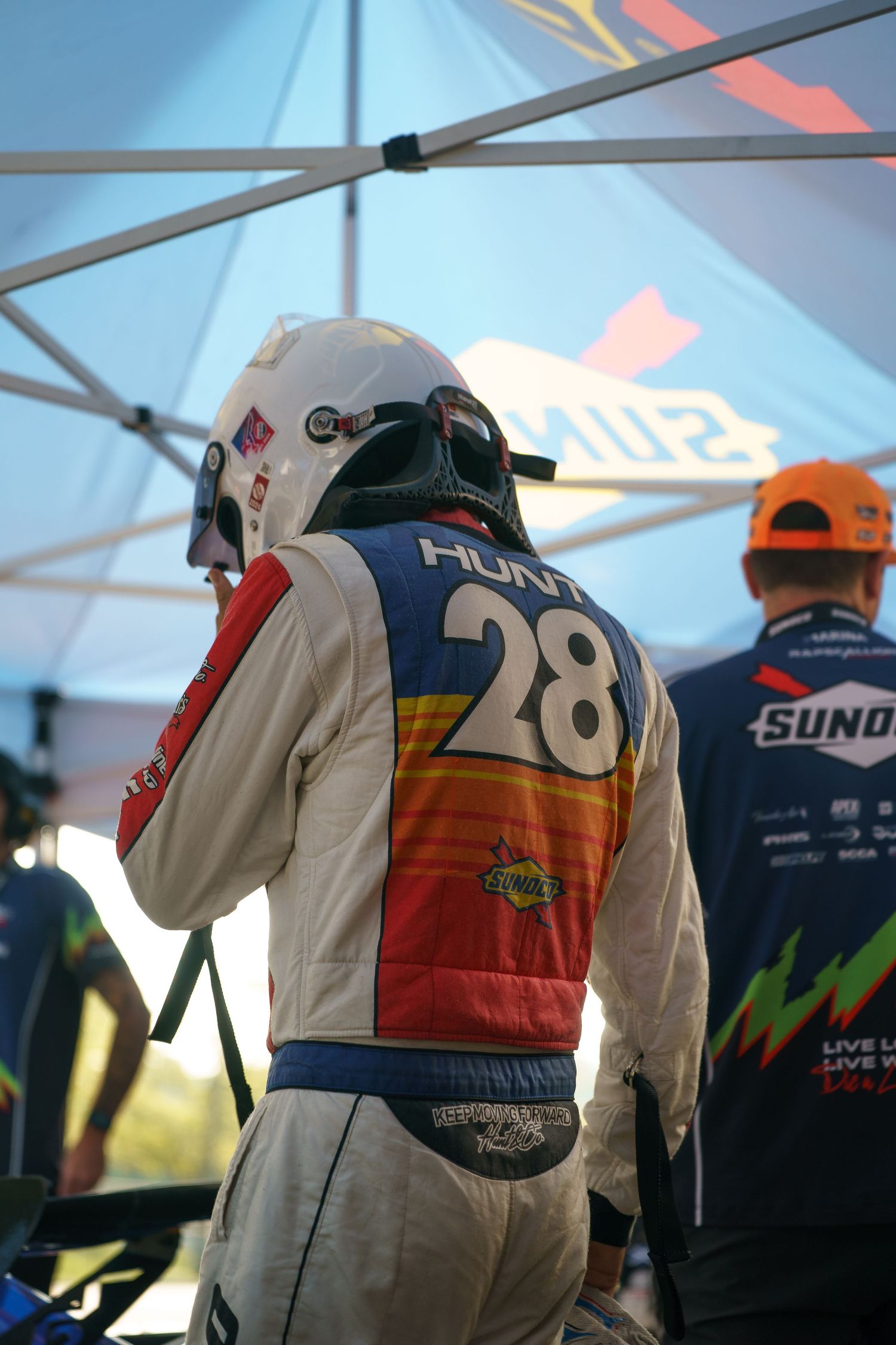 Racing driver in white firesuit with number 28 and retro-style stripes stands in pit area under blue canopy.