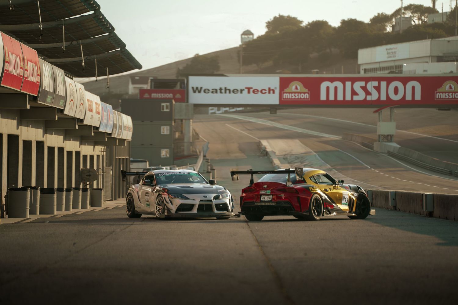 Multiple racing vehicles parked along a racetrack barrier with Mission branding visible at dusk.