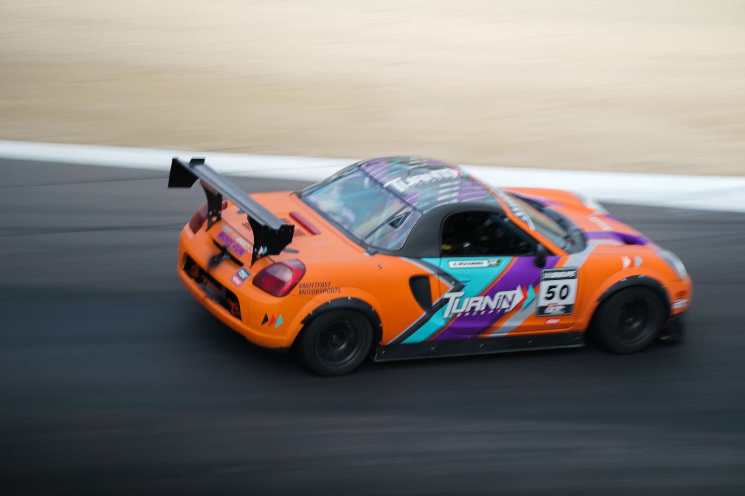 Orange race car drifting on racetrack with motion blur effect during competitive motorsport event.