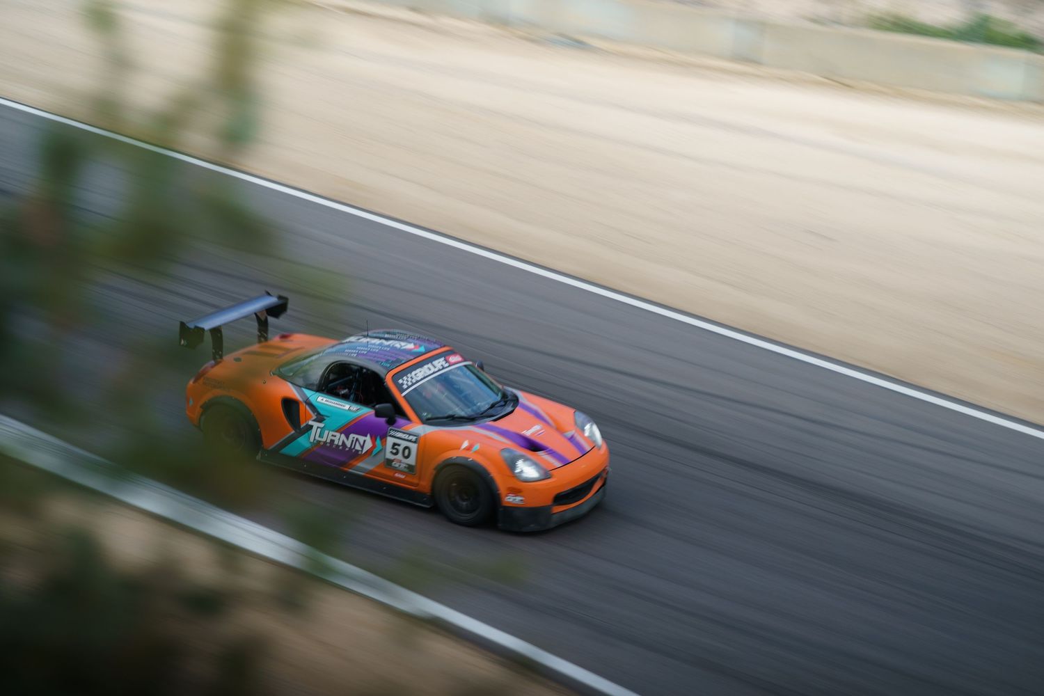 Orange and teal racing car speeds along racetrack with motion blur effect.