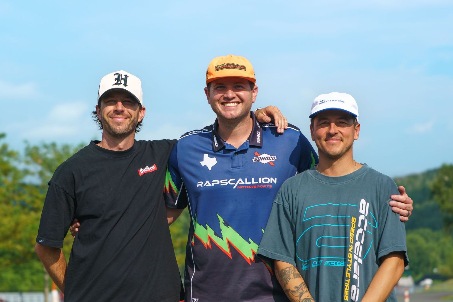 Three disc golf players pose together on a sunny day at an outdoor course.