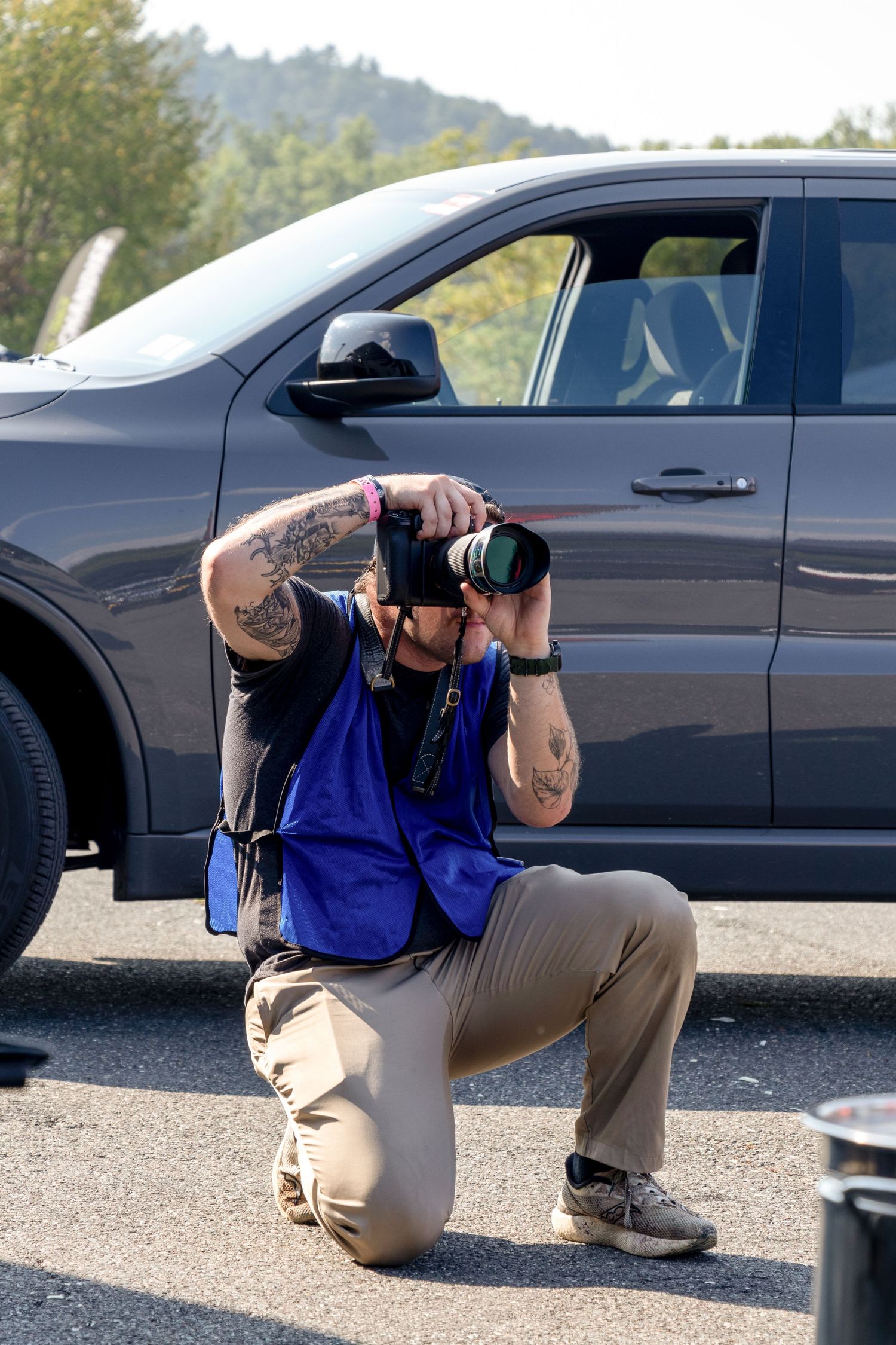 Photographer in blue vest and khaki pants demonstrates low angle shooting techniques beside a black car.