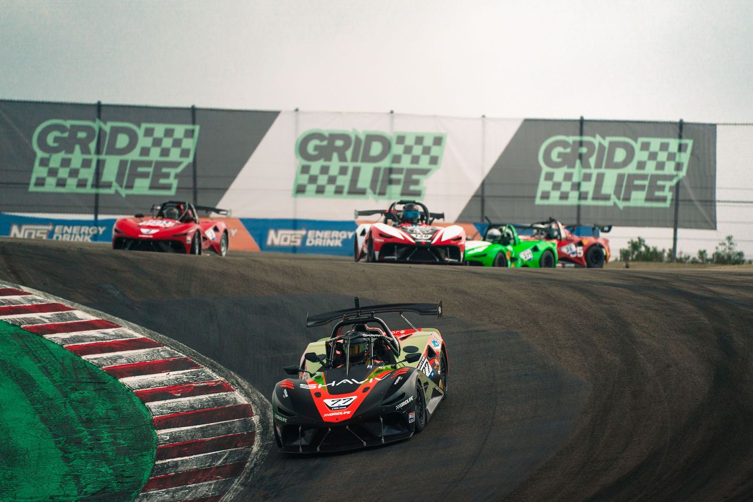 Racing cars navigate a tight corner on a Formula 1 track with green and white barriers visible in the background.