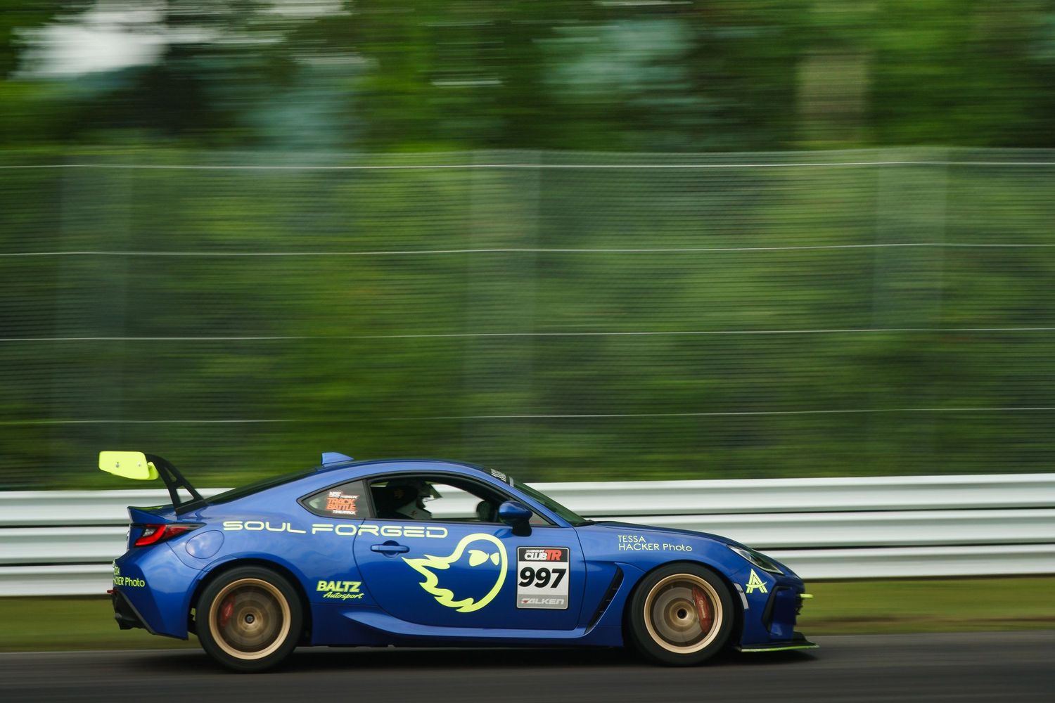 A blue sports car with racing livery speeds along a racetrack with trees and barriers visible in the background.