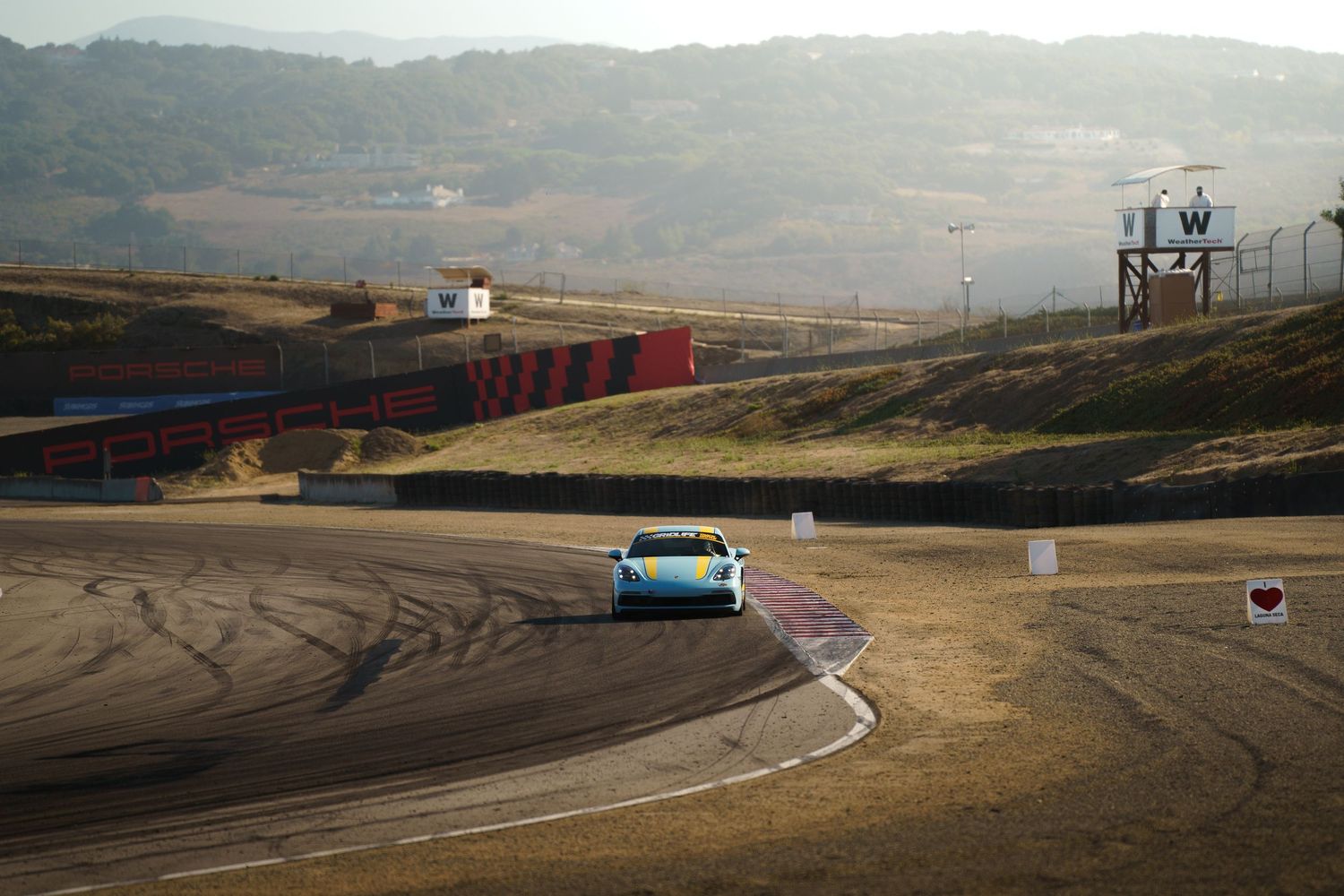 A turquoise sports car navigates a curved racetrack on a hilly terrain with tire marks visible on the asphalt.