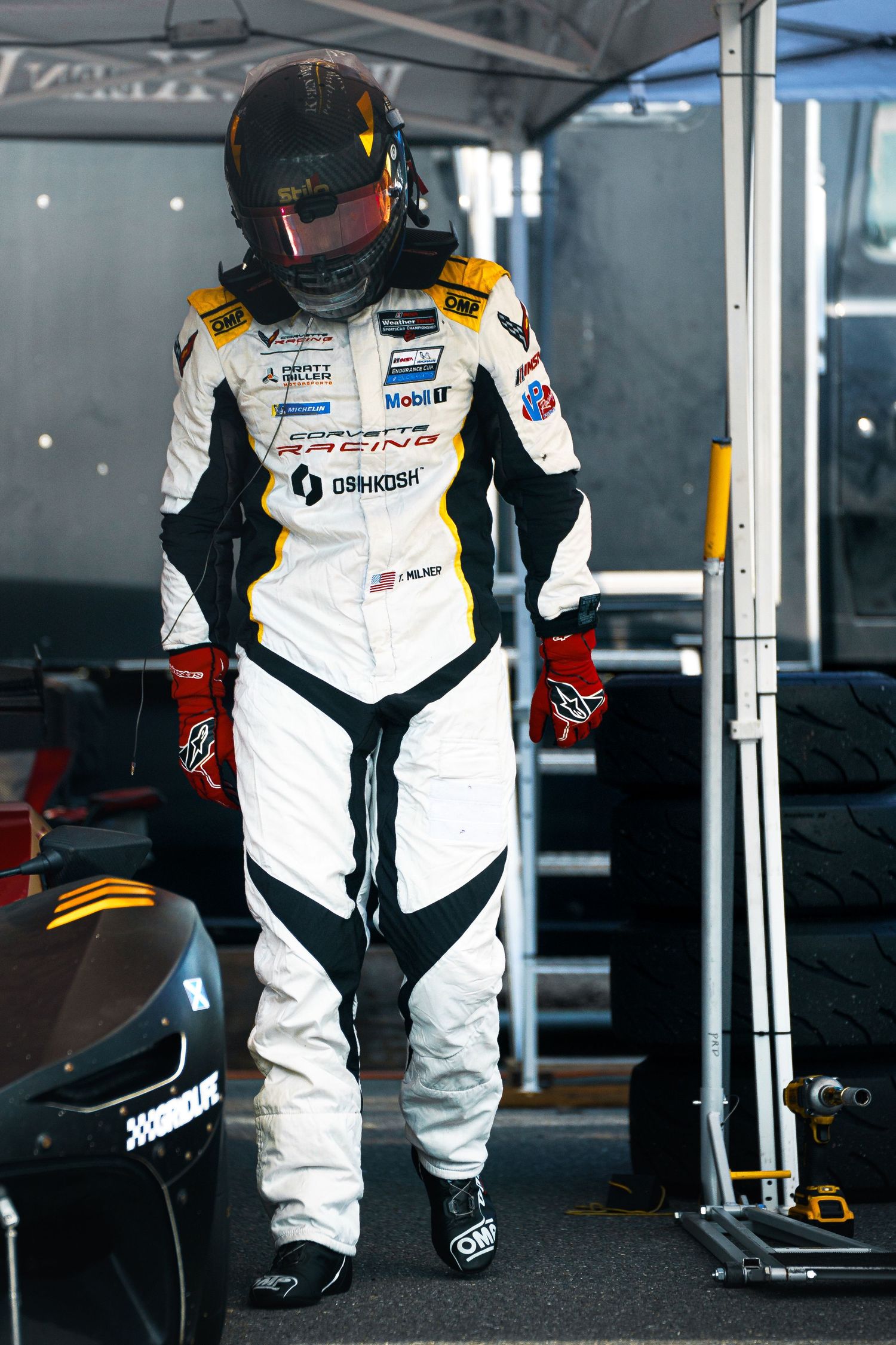 Race driver in white protective racing suit and gear standing in pit area.