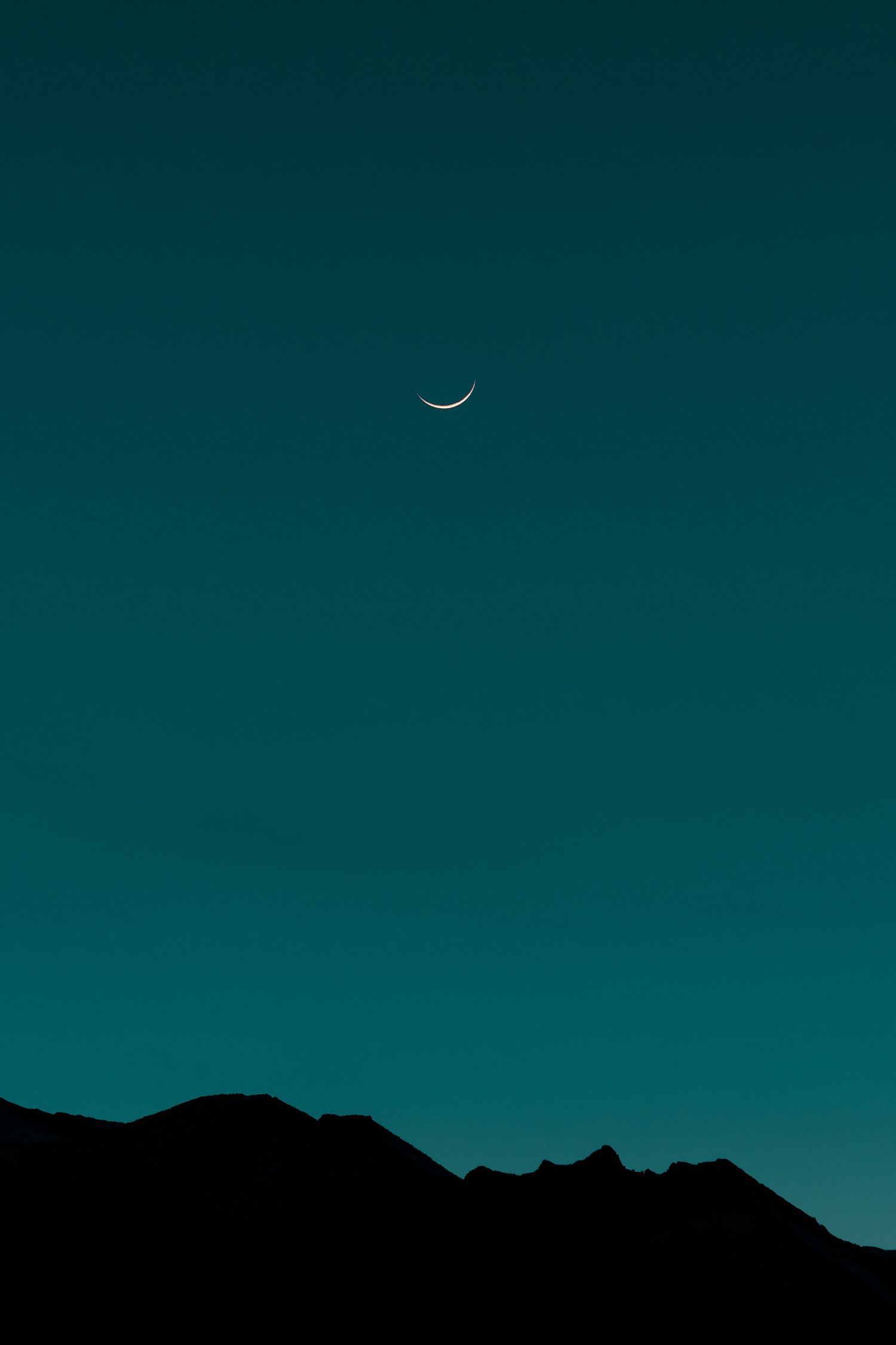 Thin crescent moon glows in a clear teal sky above the dark silhouette of a mountain ridge.