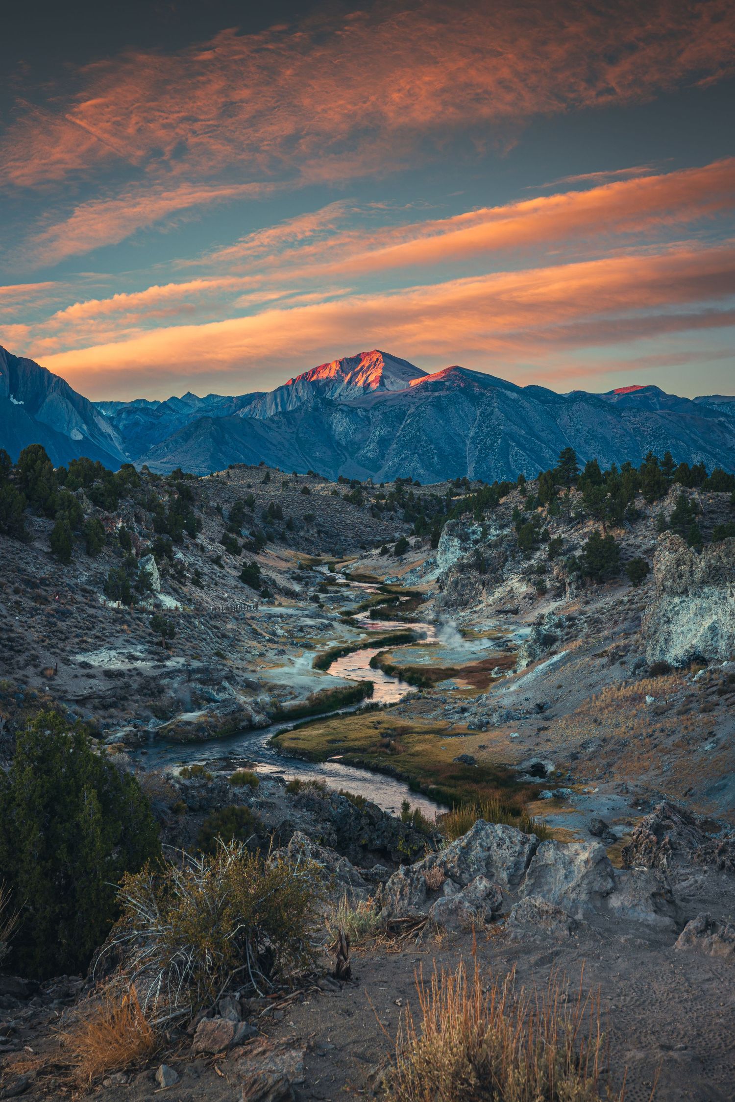 Winding creek flows through a rugged valley at sunset with a snow-capped mountain peak glowing orange.
