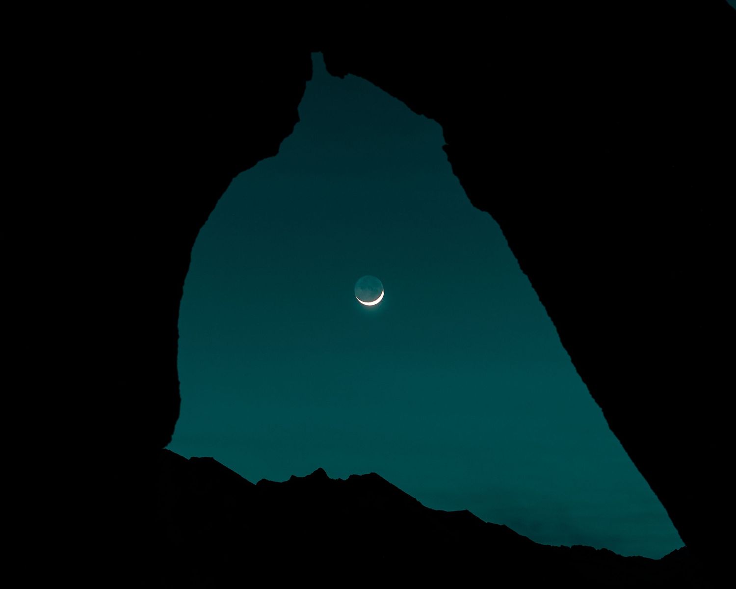 Crescent moon visible through a dark silhouetted rock formation against a deep teal night sky.