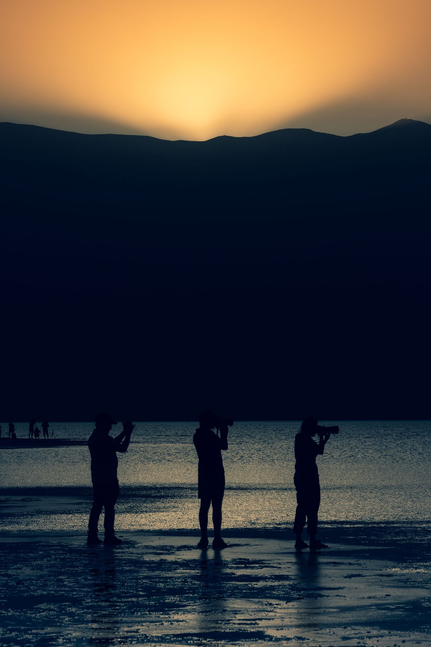Three silhouetted photographers stand in shallow water capturing a vibrant golden sunset behind dark mountains.