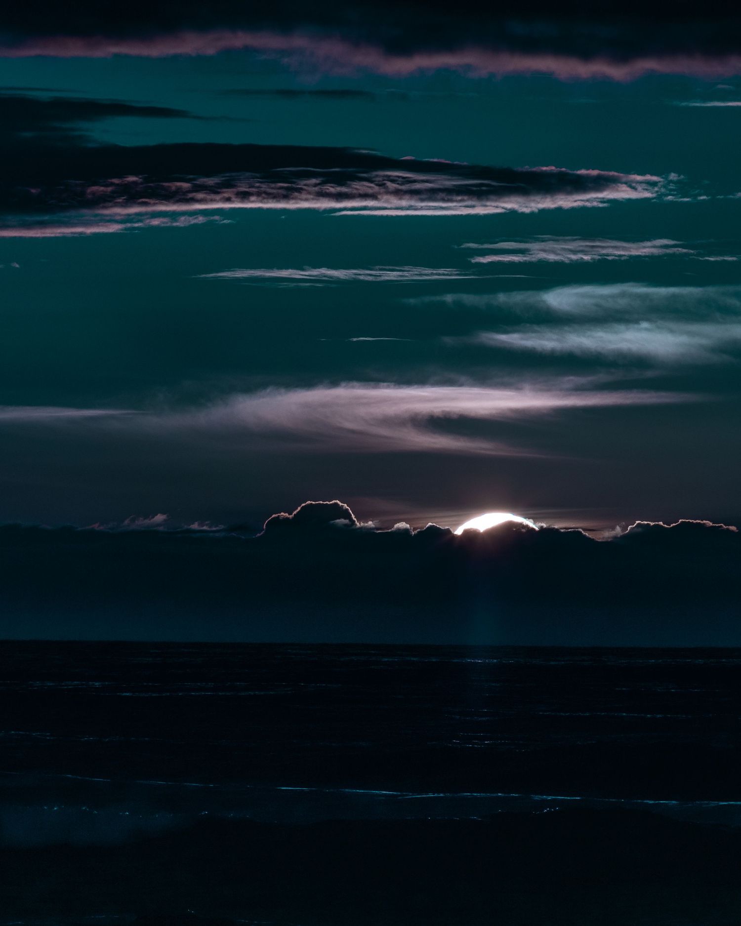 Dark dramatic seascape with glowing sun peeking through teal storm clouds over a calm ocean.