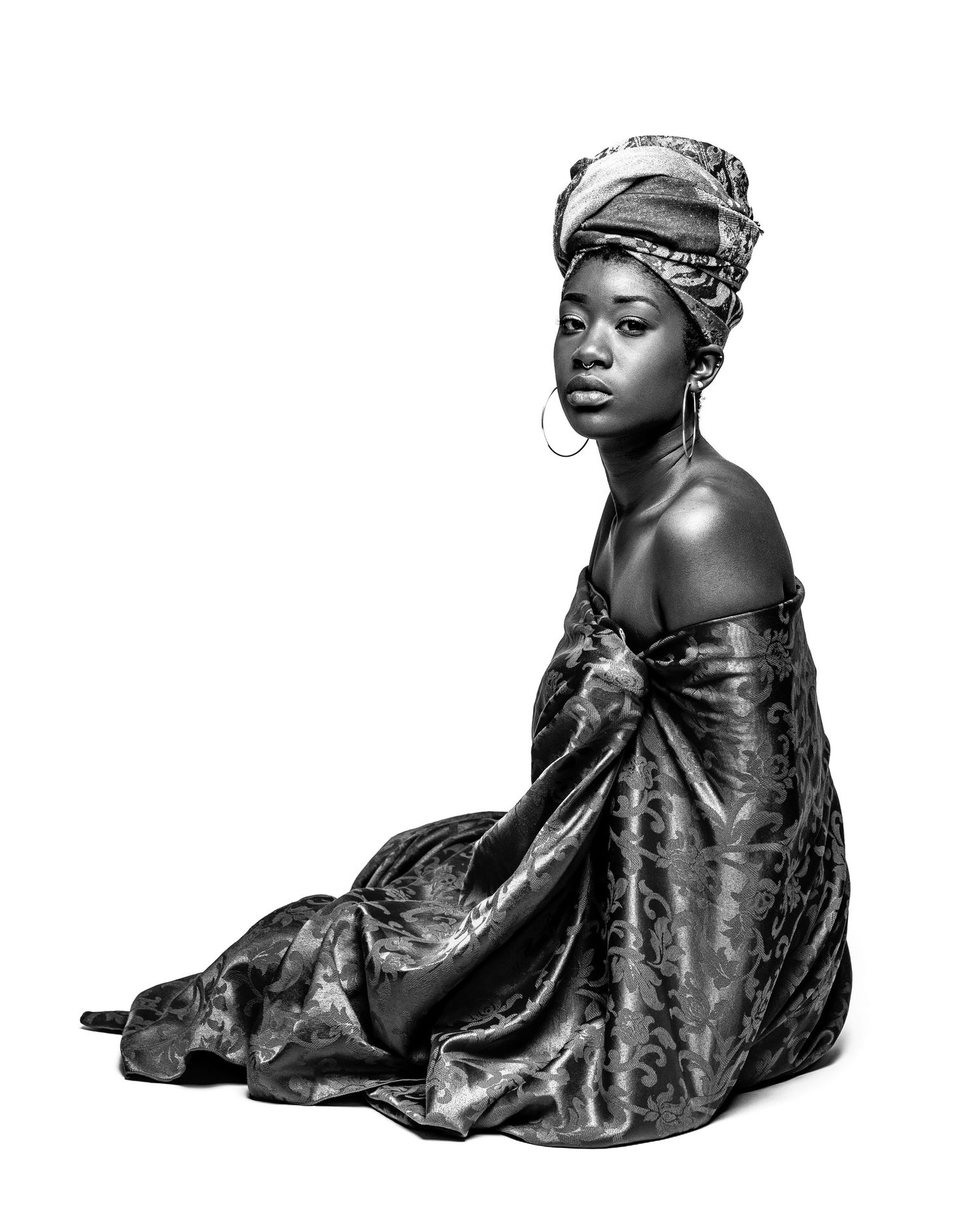 Black and white portrait of a woman in an elegant dress and head wrap, sitting gracefully on a white background.