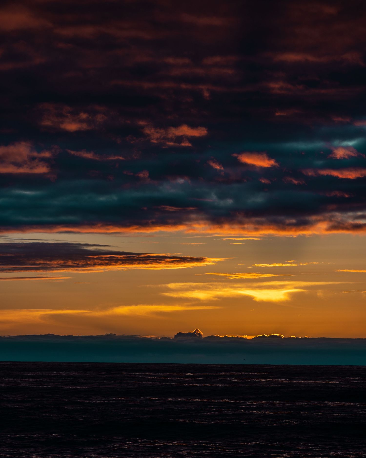Dramatic ocean sunset with dark storm clouds illuminated by golden orange light on the horizon.