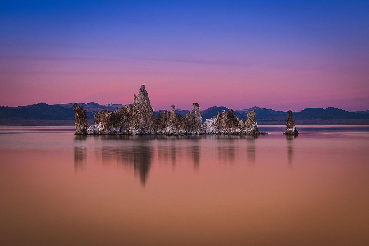 Tufa rock formations reflected in calm pink and purple waters of Mono Lake at sunset.