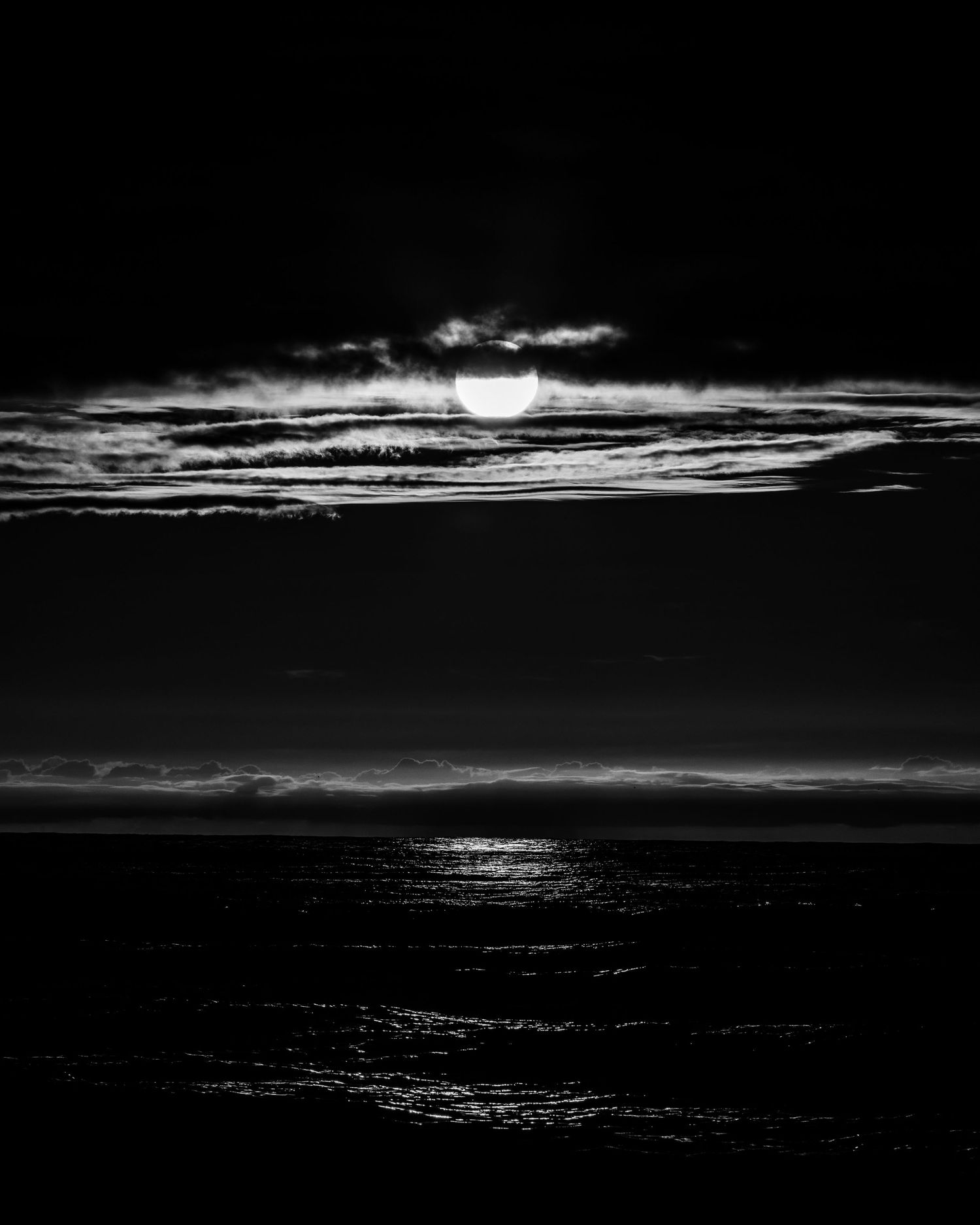 Black and white moonrise over ocean with dramatic clouds and moonlight reflecting on dark water.