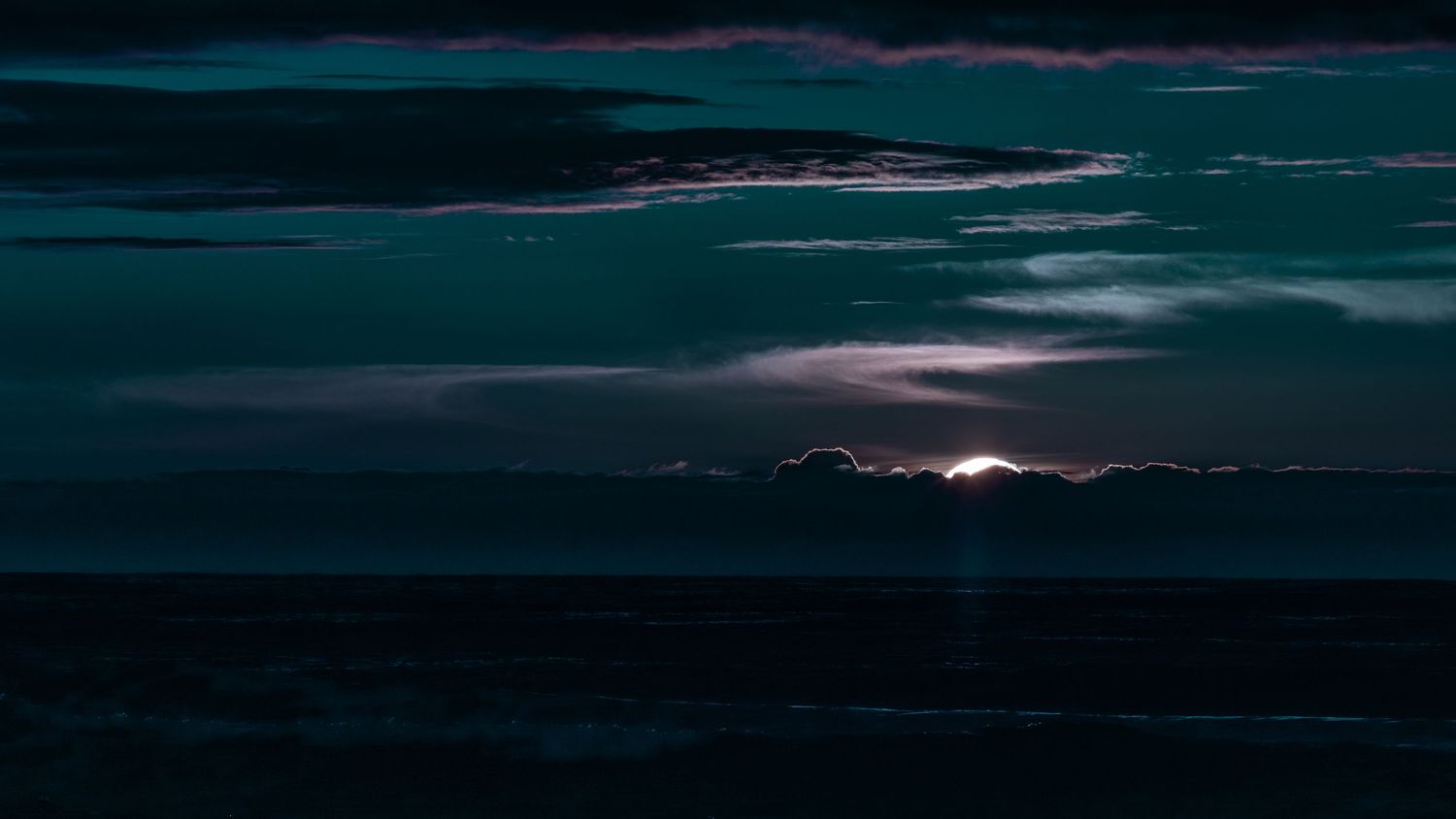 Dark moody ocean horizon with glowing moonlight breaking through dramatic storm clouds.