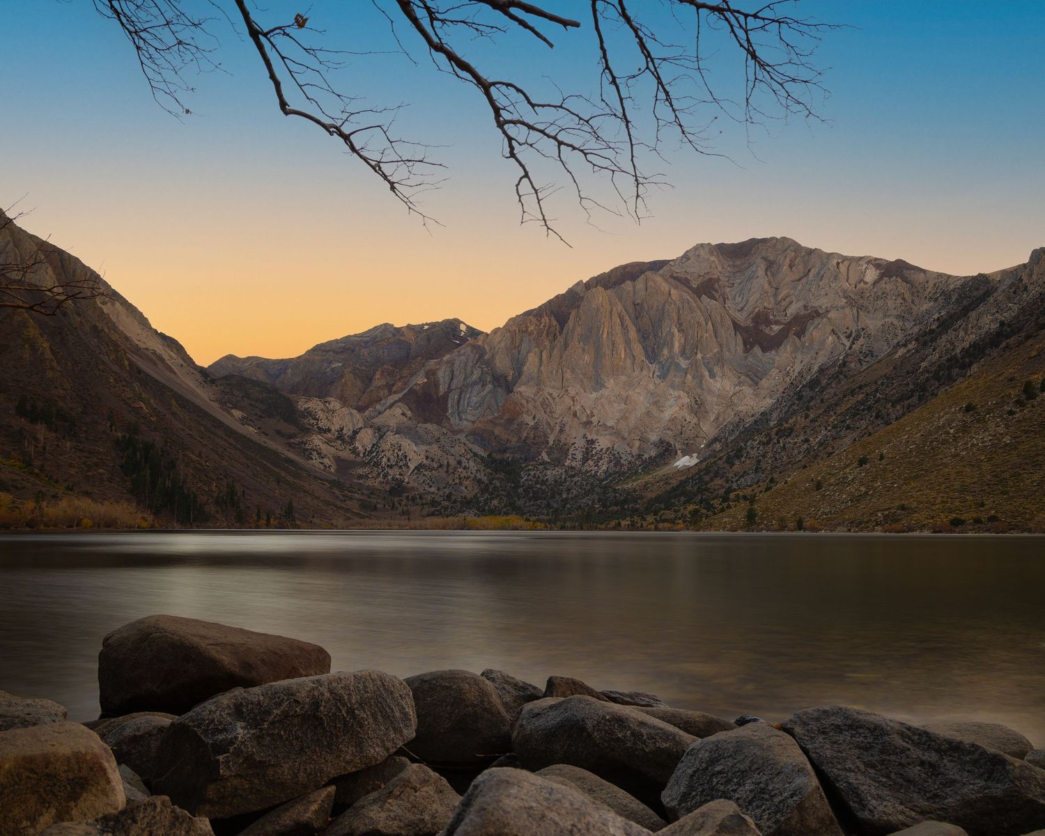 Tranquil mountain lake at sunset with rocky shoreline, bare tree branches, and towering cliffs in background.