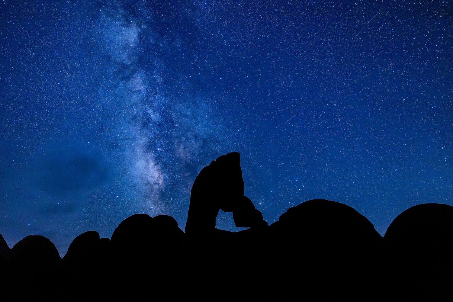 Silhouette of a rock arch against a stunning Milky Way galaxy in a deep blue starry night sky.