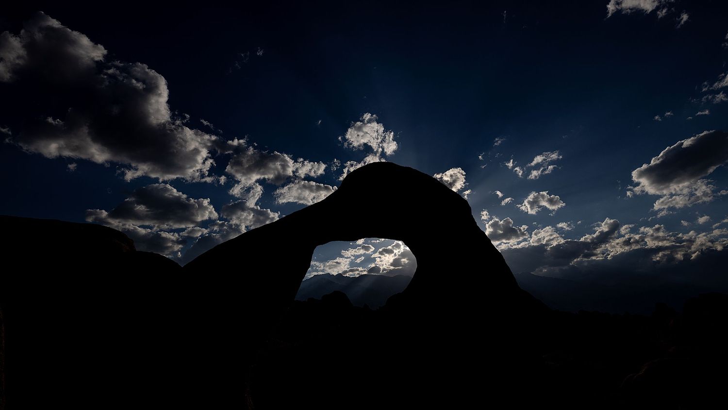 Dramatic silhouette of a natural rock arch against a moody blue night sky with scattered clouds.
