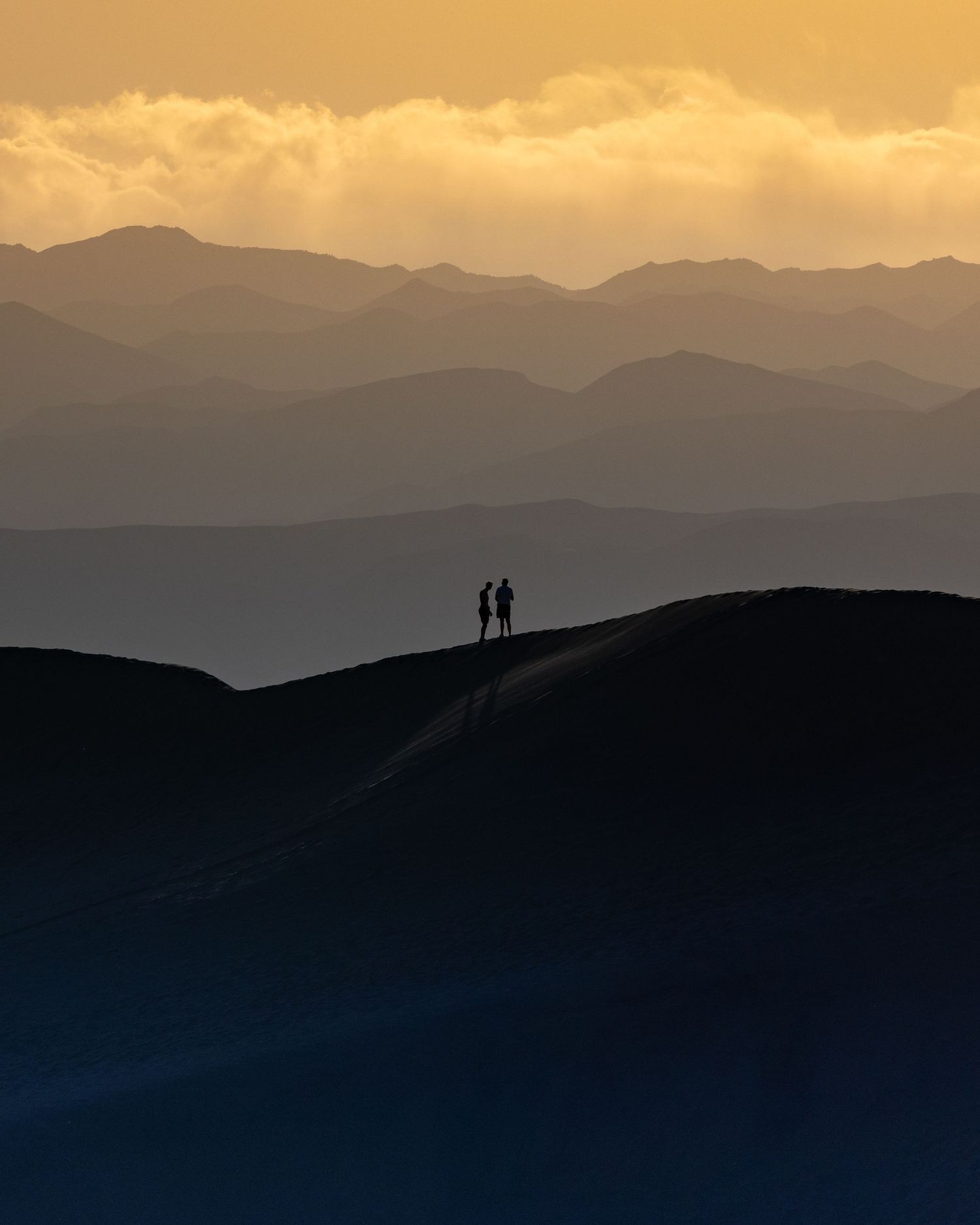 Two silhouetted figures stand on a hilltop at sunset with layered mountain ranges behind.