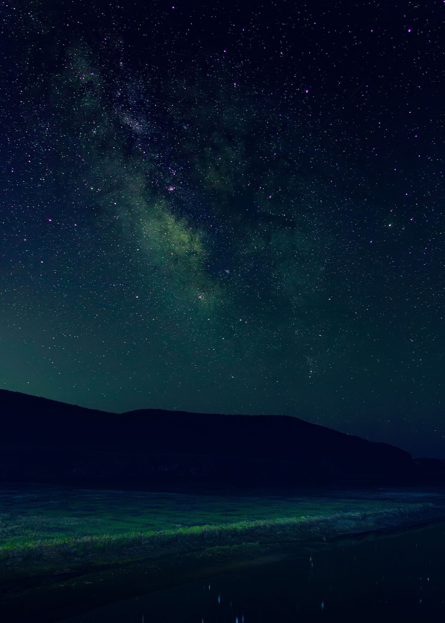 Milky Way galaxy glowing green over dark silhouetted hills reflected in calm water.