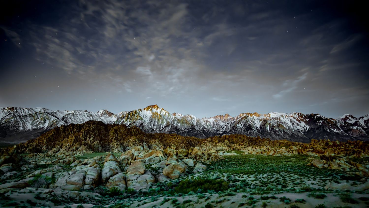 Snow-capped mountain range rising above golden foothills and white boulders under a cloudy sky.