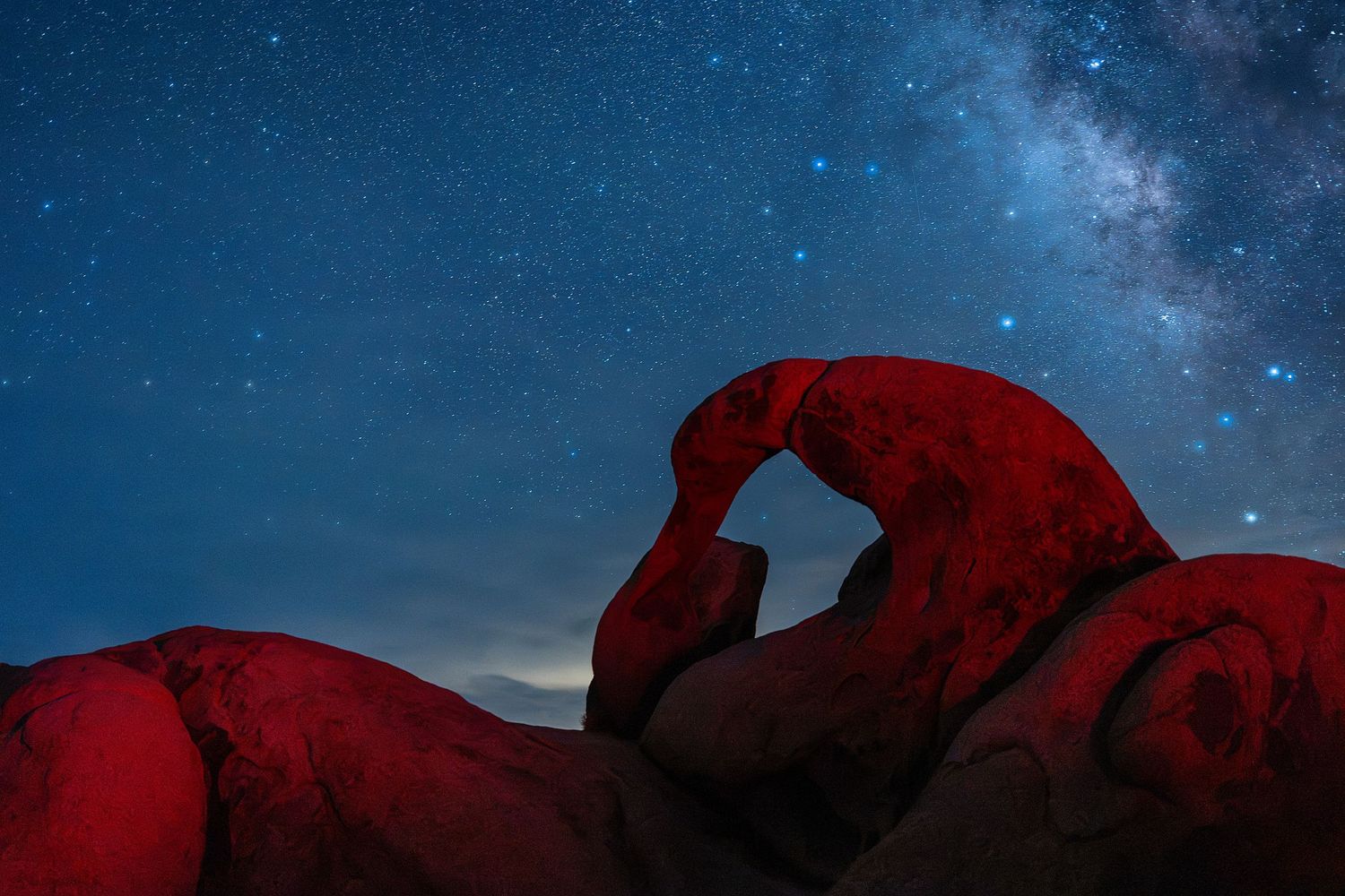Red-lit rock arch silhouetted against a stunning Milky Way night sky in the desert wilderness.