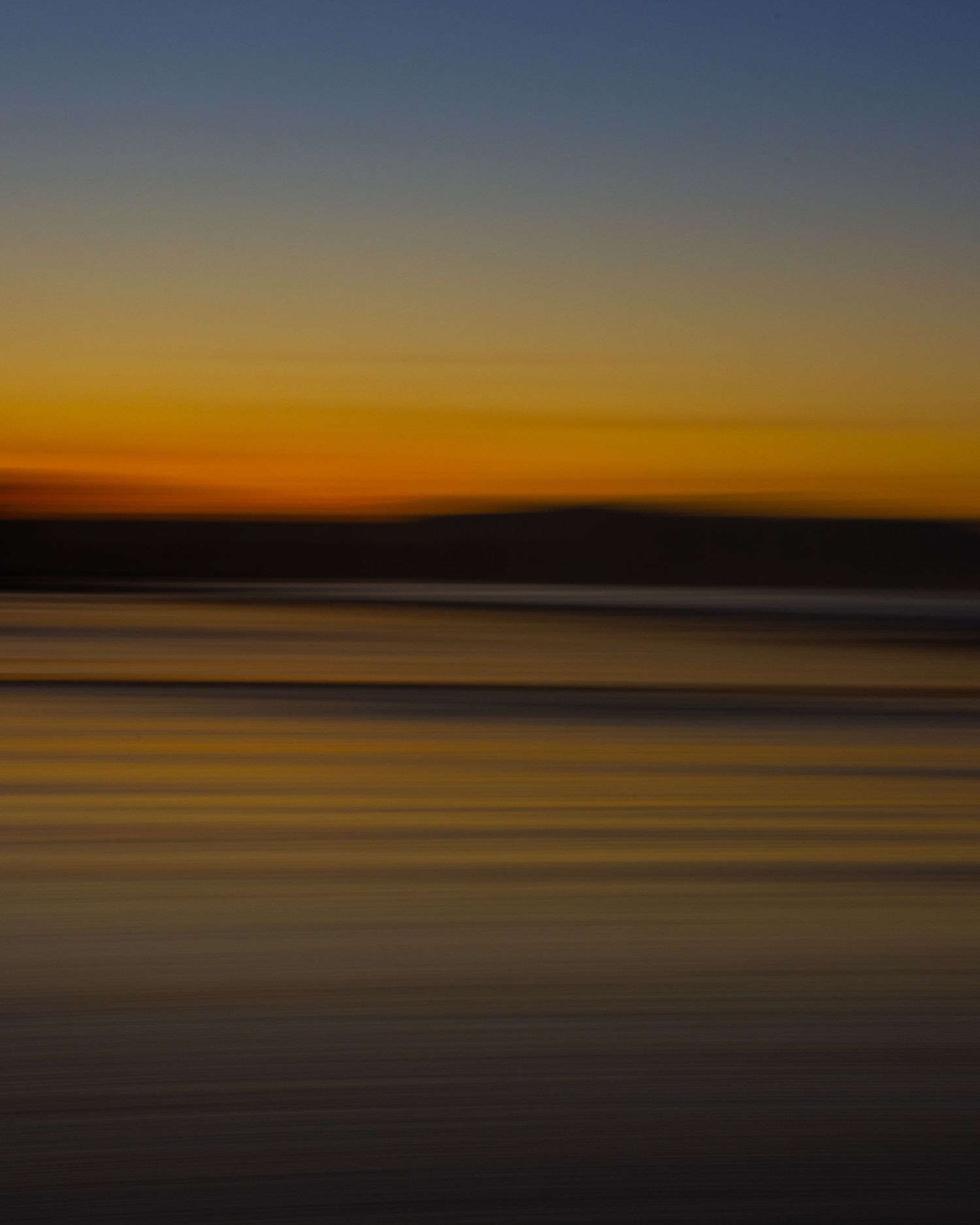 Blurred coastal sunset with glowing orange horizon, dark landmass silhouette, and calm water.