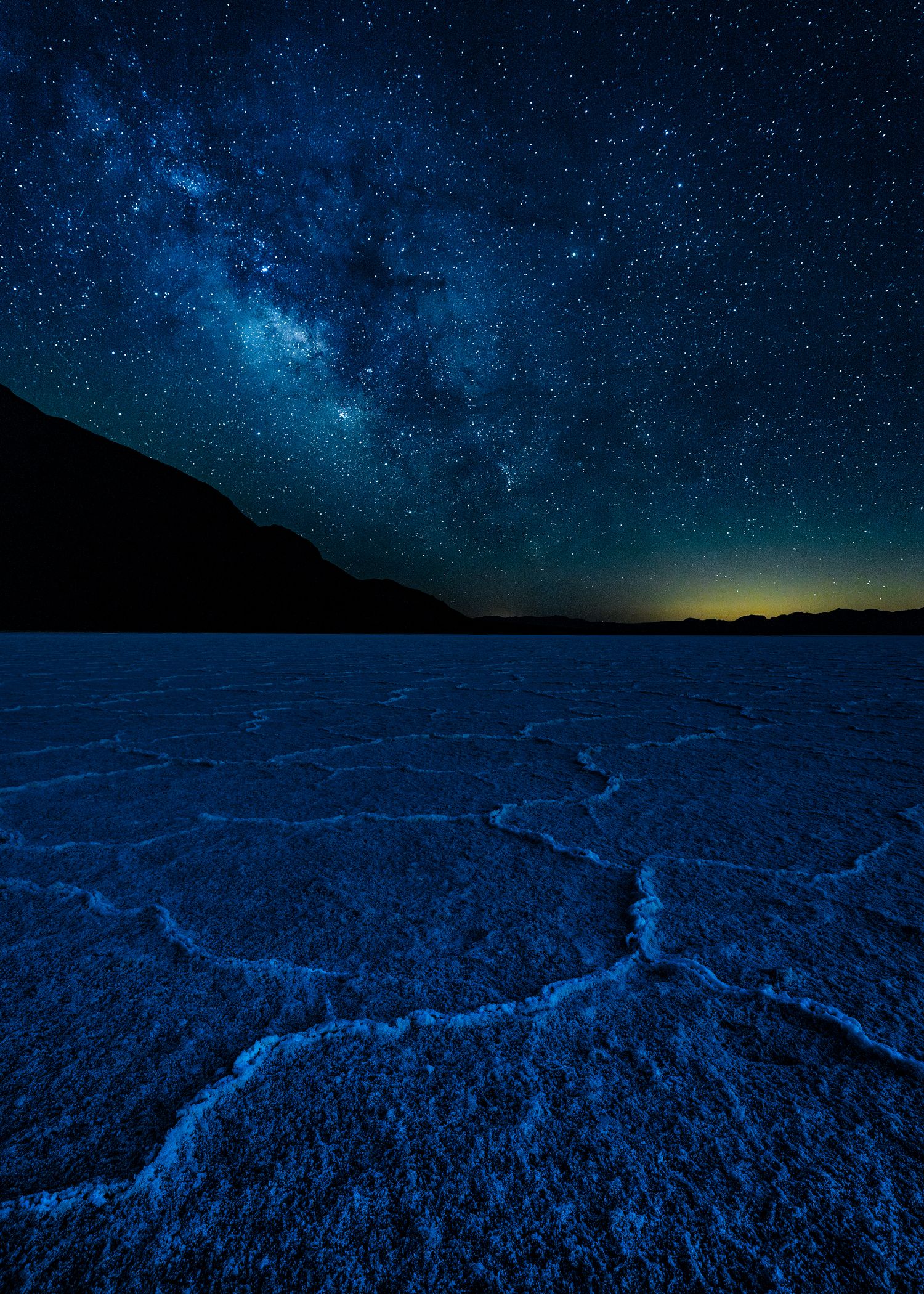 Salt flats under a stunning Milky Way night sky with stars and mountains silhouetted on the horizon.