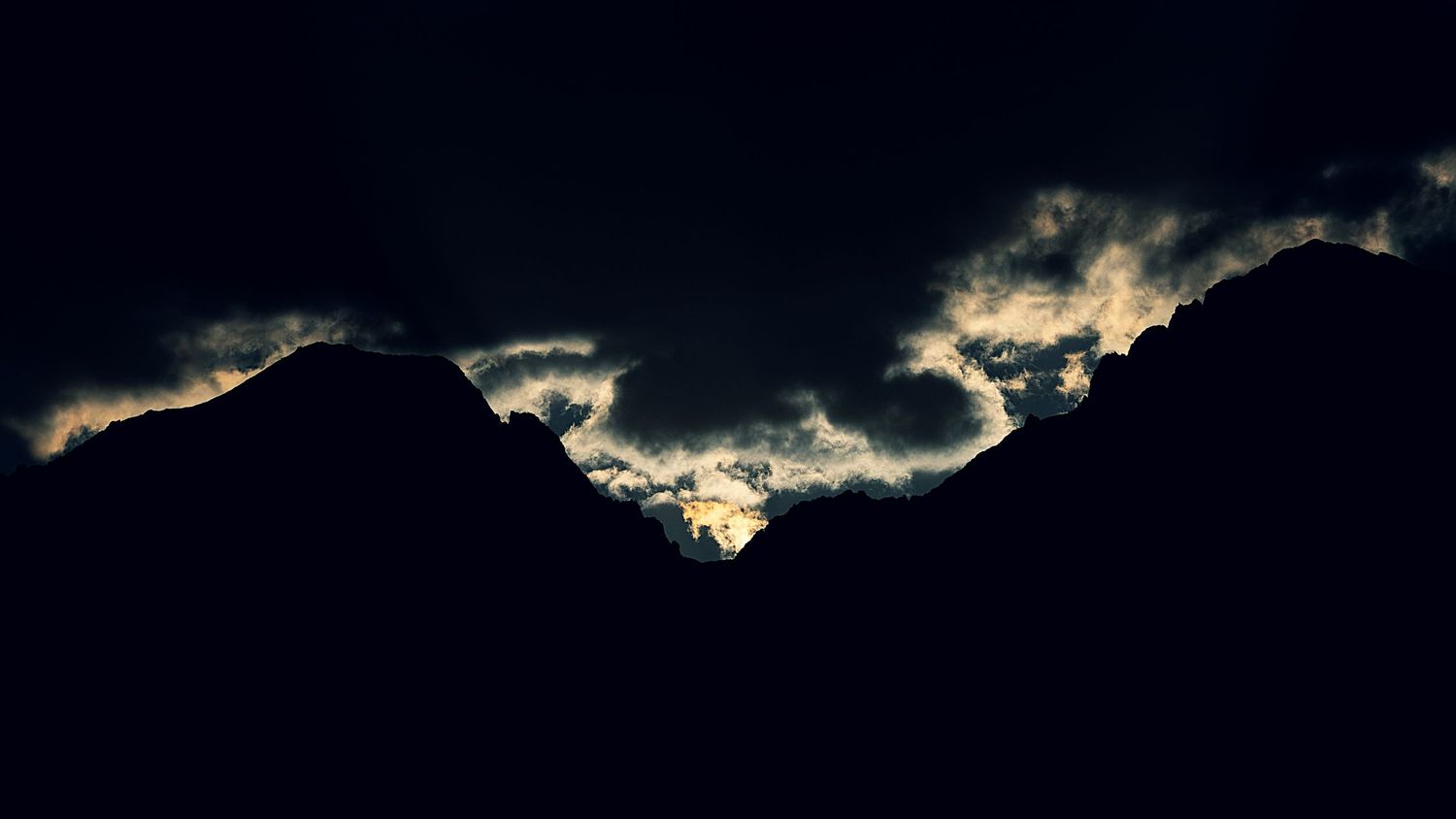 Dark silhouette of mountain peaks against a dramatic sky with glowing clouds at dusk.