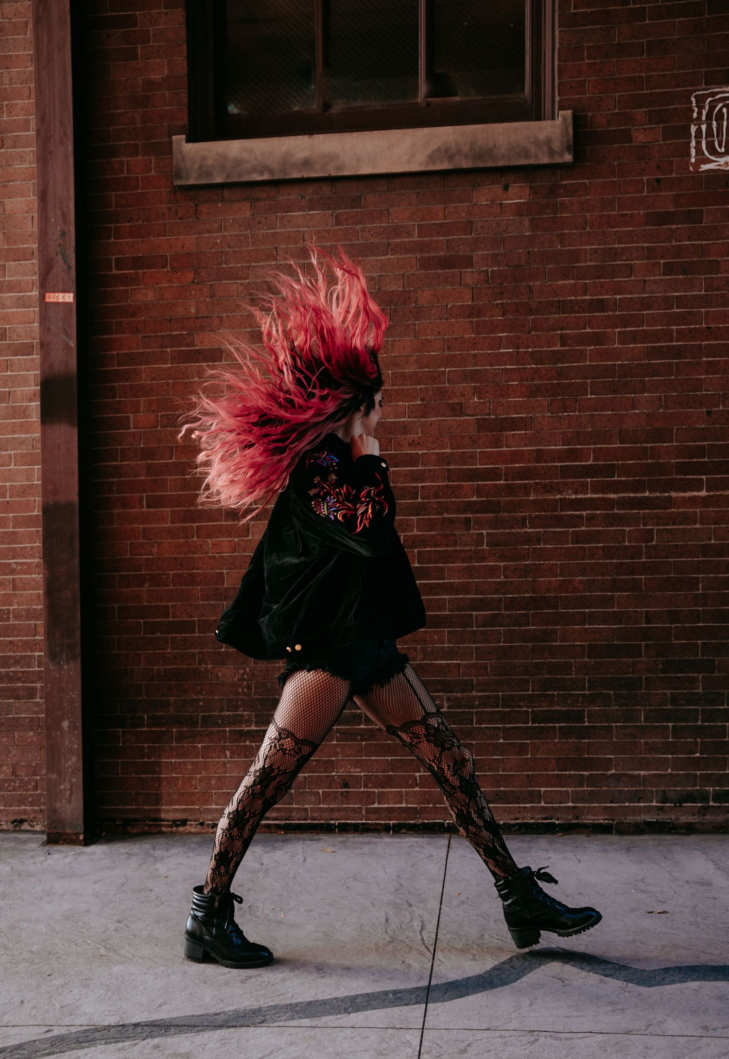 Woman with vibrant pink hair tossing hair while walking past a red brick building.