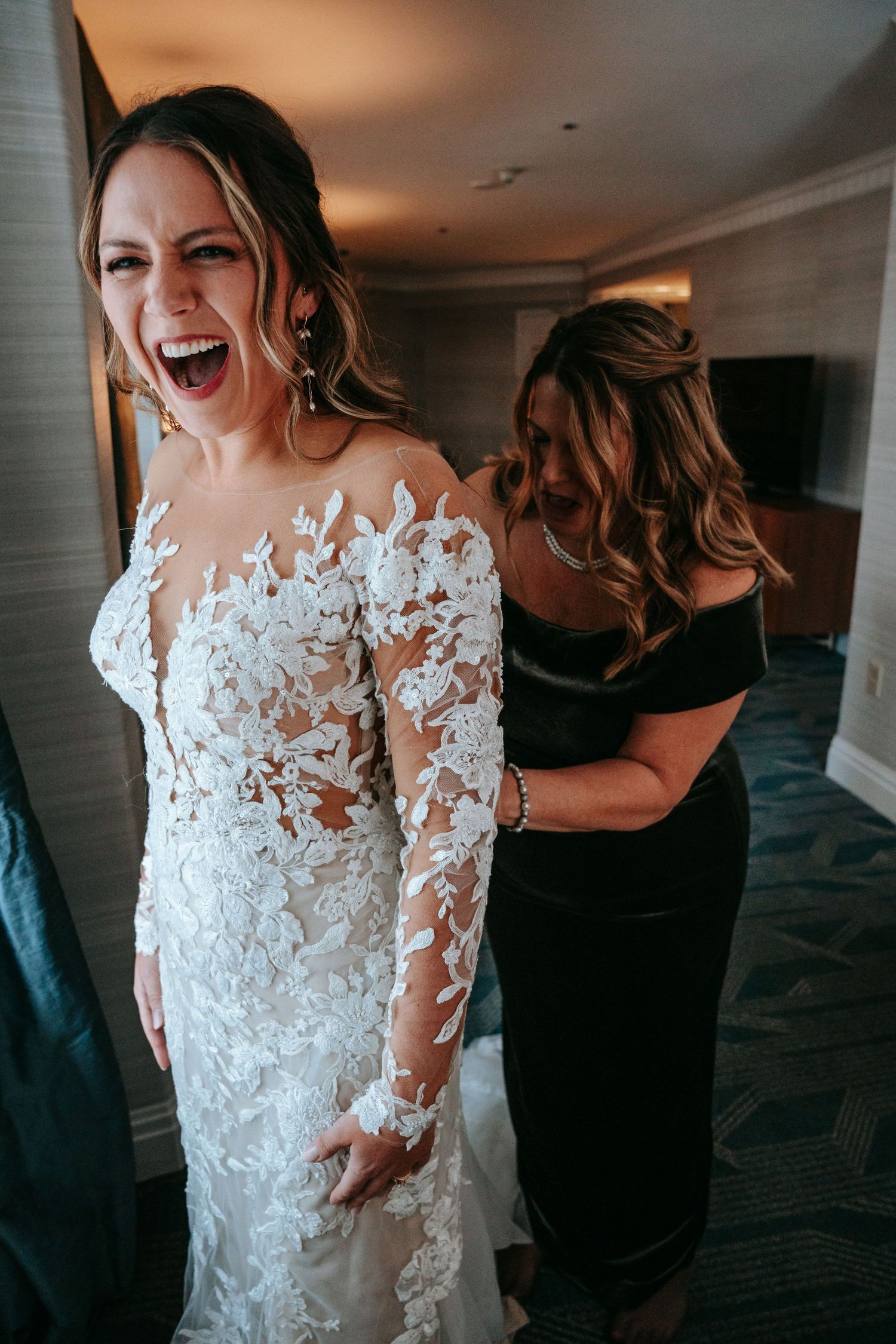 Bride laughing joyfully as bridesmaid buttons up her long-sleeve lace wedding gown in hotel room.