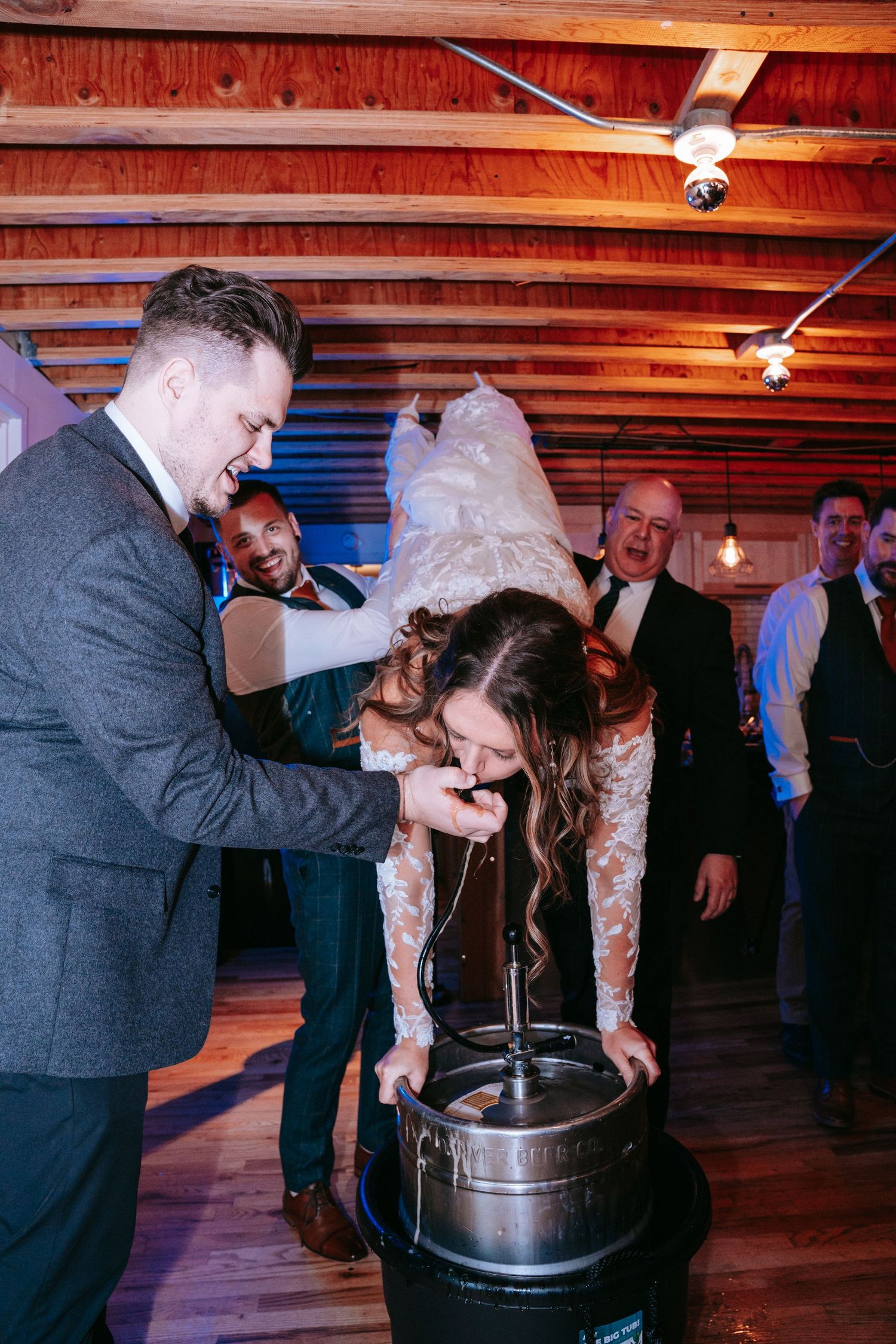 Bride doing a keg stand assisted by groom at a rustic wood-ceiling wedding reception.