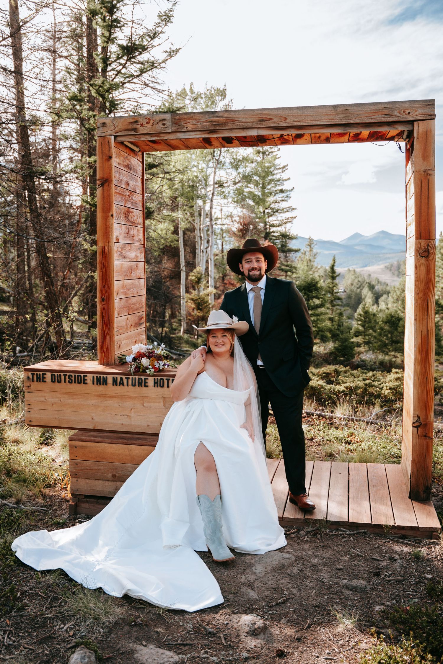 Bride in white gown and hat sits with groom in dark suit and cowboy hat at outdoor wedding at The Outside Inn Nature Hotel.