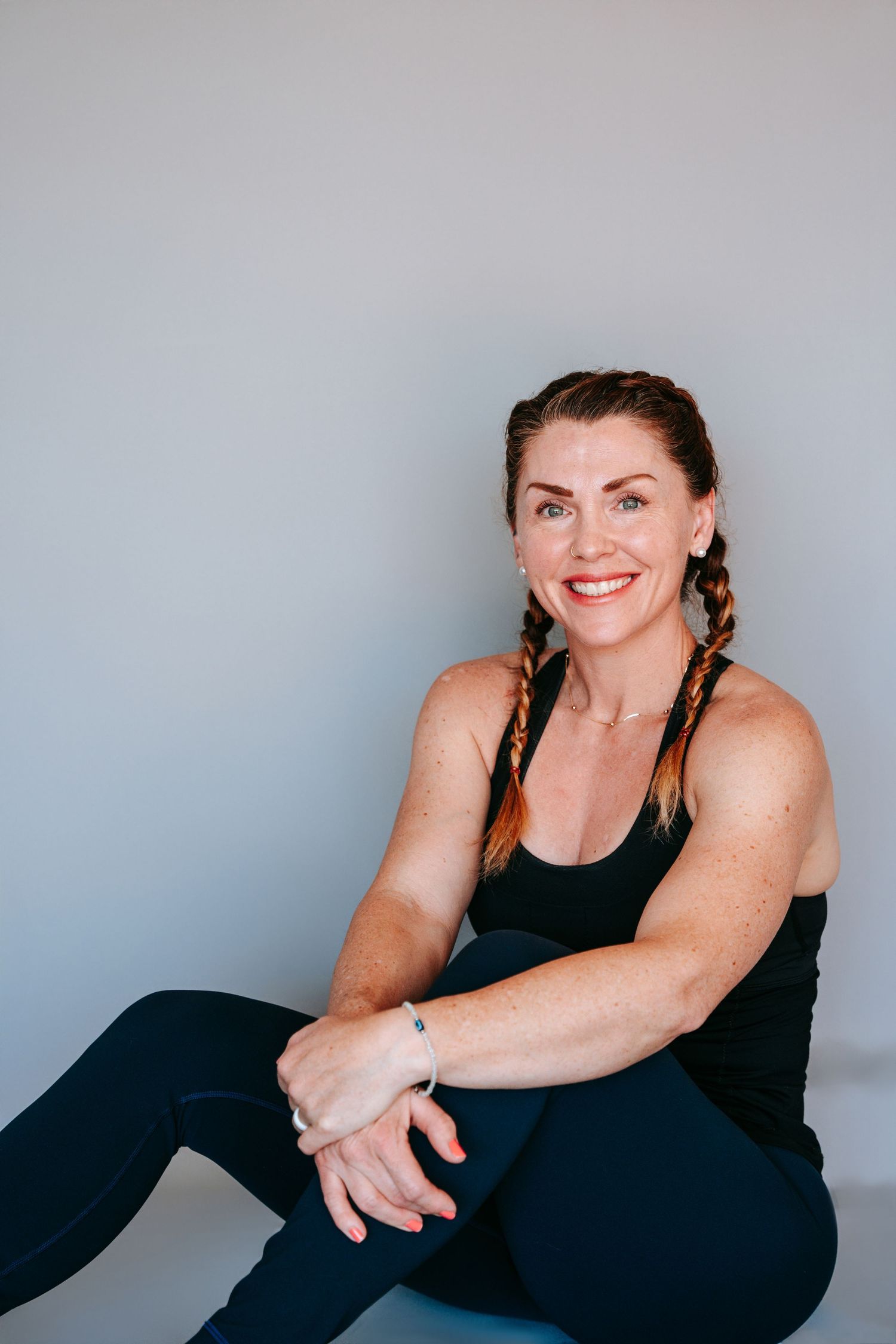 Fit woman with braided hair in black athletic wear sitting relaxed against light gray wall.