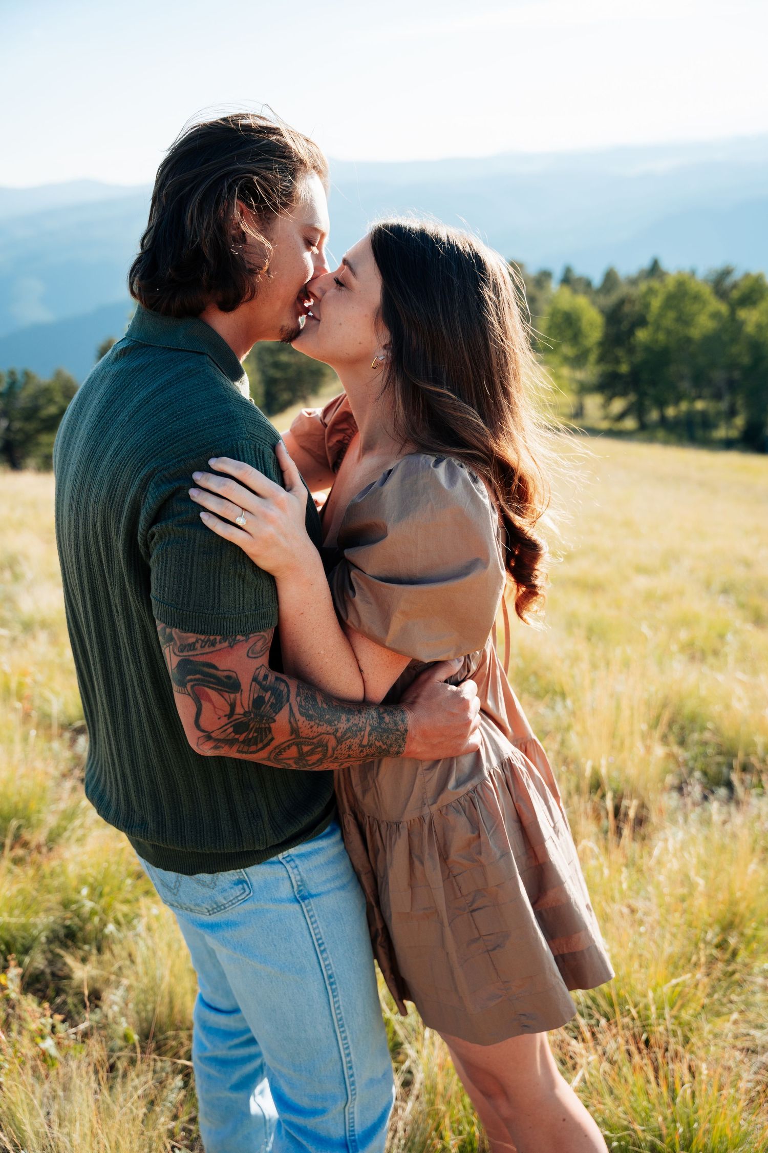 Engaged couple sharing a romantic kiss in a sunlit mountain meadow, man wearing green sweater with tattoos.