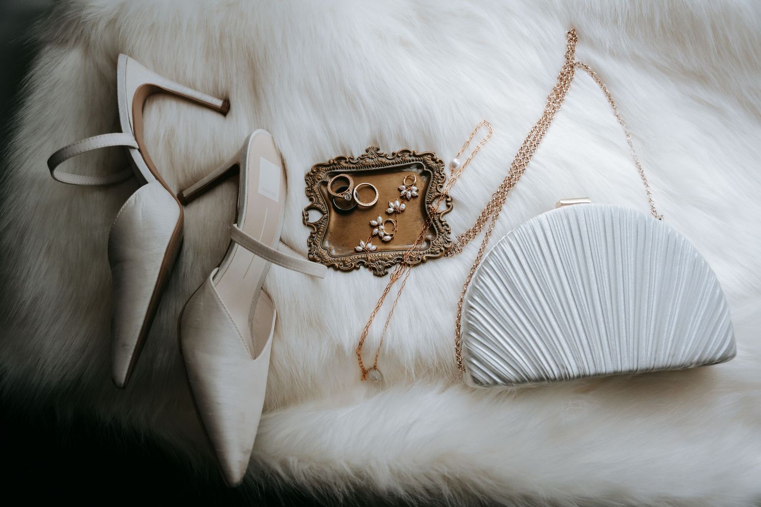 Elegant wedding accessories including nude heels, white clutch, and gold jewelry on fur surface.