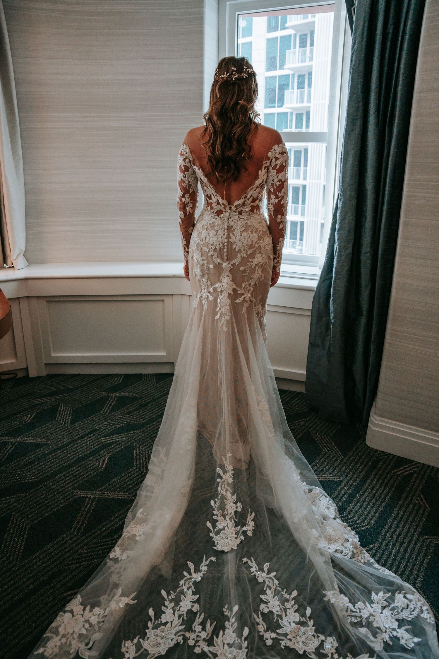 Bride in a stunning long-sleeve lace wedding gown with floral train standing by window.