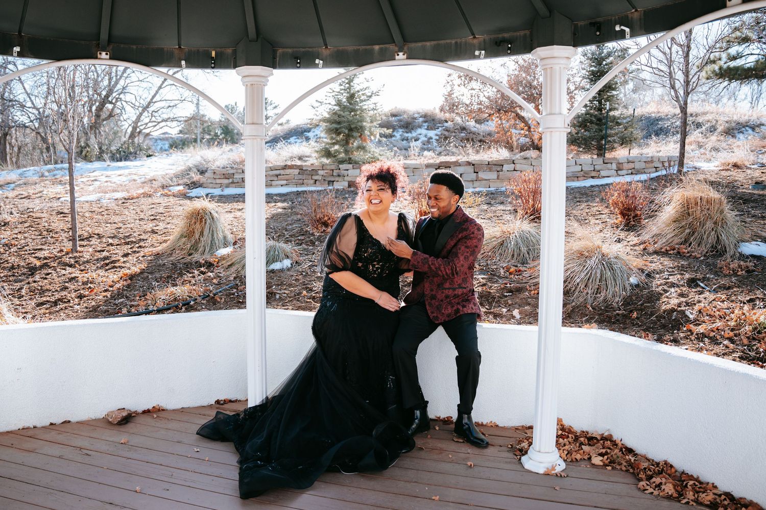 Couple poses in a white gazebo wearing black wedding attire during a winter outdoor ceremony.